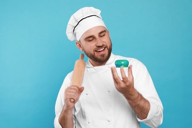 Happy professional confectioner in uniform with delicious cake and rolling pin on light blue background
