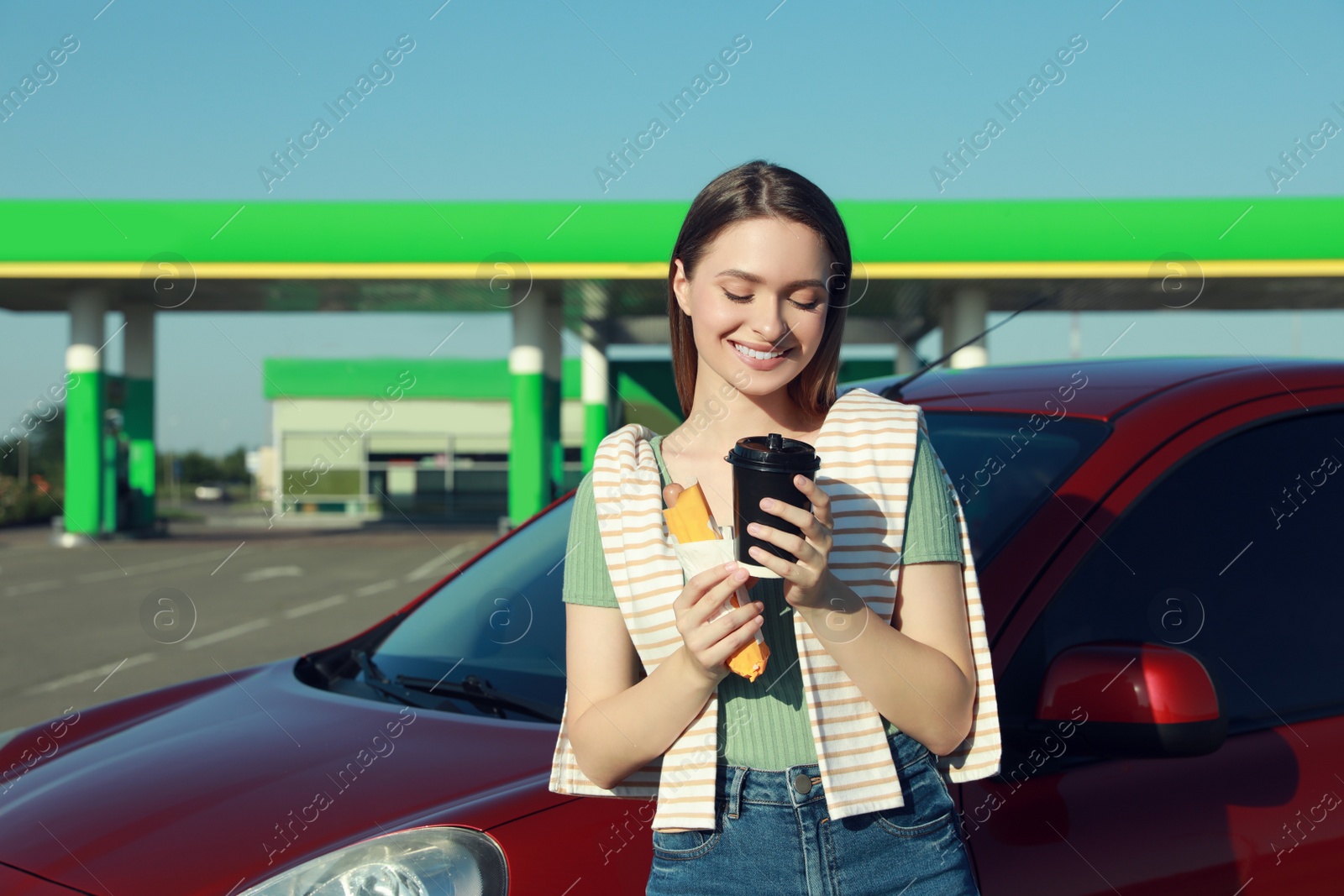 Photo of Beautiful young woman with coffee and hot dog near car at gas station