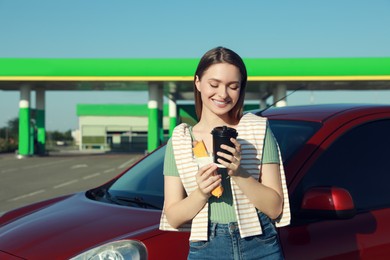 Photo of Beautiful young woman with coffee and hot dog near car at gas station