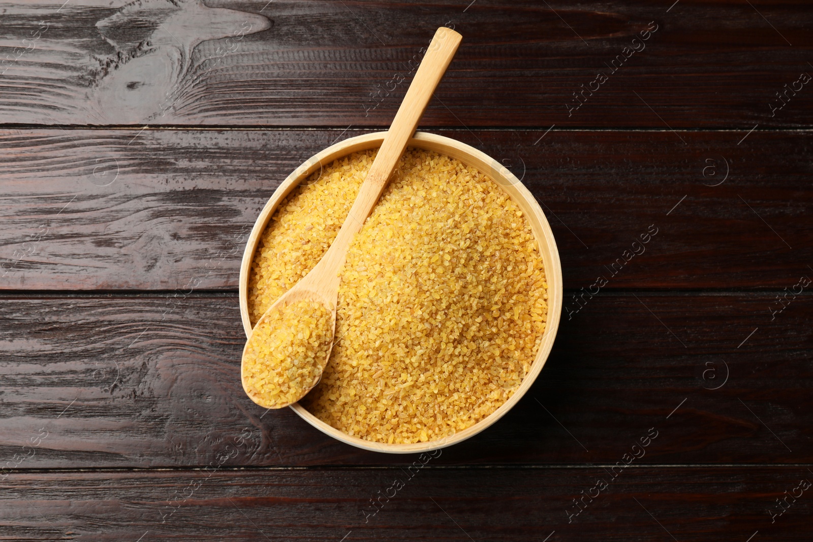 Photo of Bowl and spoon with raw bulgur on wooden table, top view