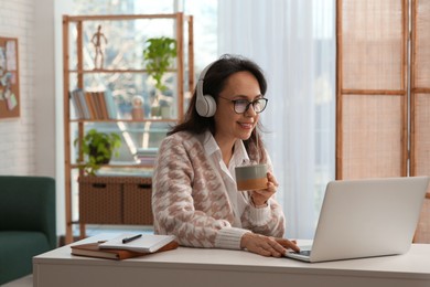 Photo of Woman with modern laptop and headphones drinking tea while learning at table indoors