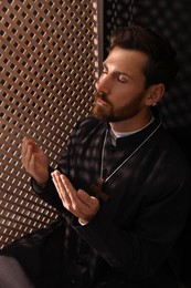 Catholic priest in cassock praying to God in confessional booth