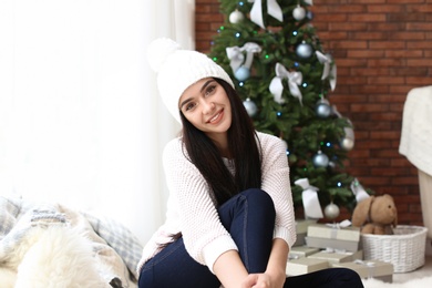 Beautiful young woman in hat near Christmas tree at home