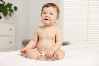 Photo of Cute little baby with moisturizing cream on face sitting on bed at home