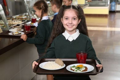 Photo of Cute girl holding tray with healthy food in school canteen