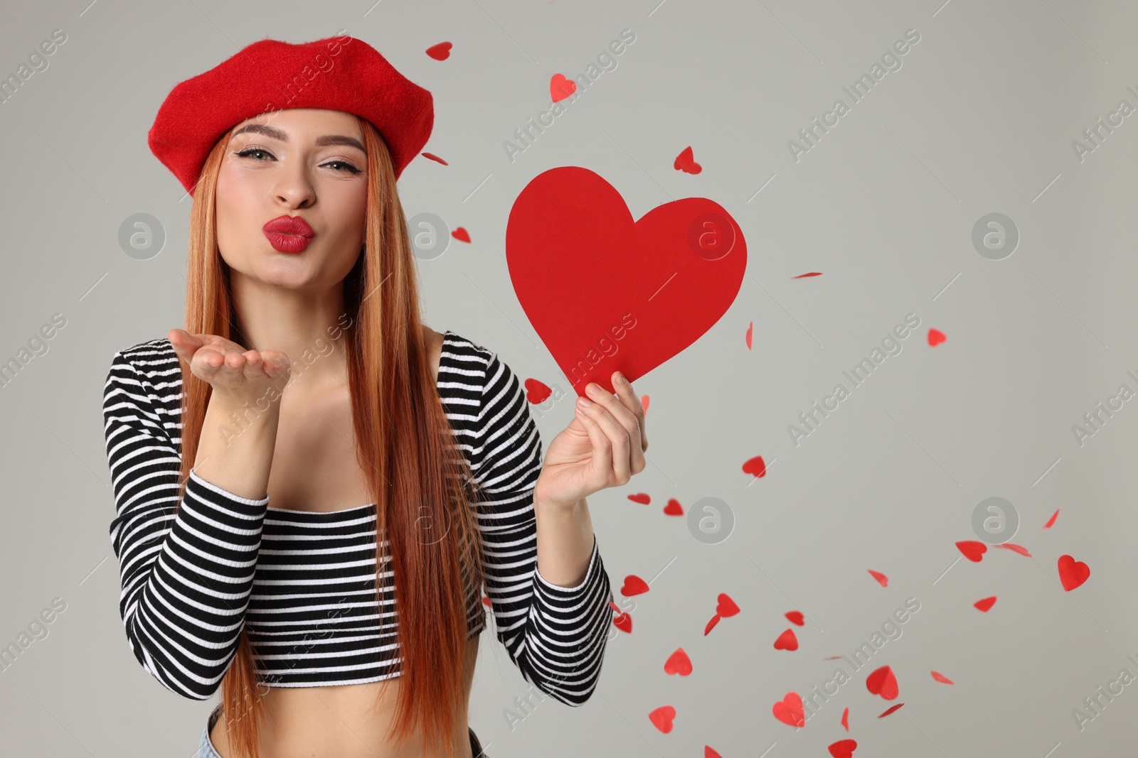 Photo of Young woman with paper heart blowing kiss under confetti on light grey background, space for text