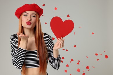 Young woman with paper heart blowing kiss under confetti on light grey background, space for text