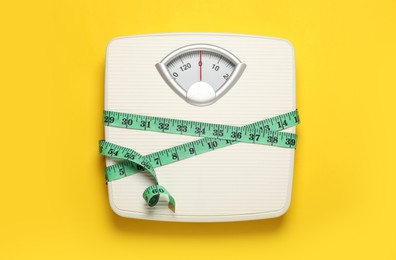 Photo of Weigh scales tied with measuring tape on yellow background, top view. Overweight concept