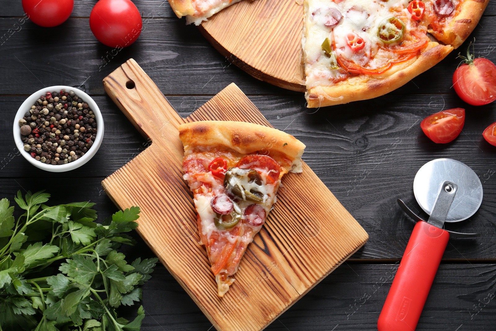 Photo of Delicious pizza Diablo, tomatoes, parsley, peppercorns and cutter on black wooden table, flat lay