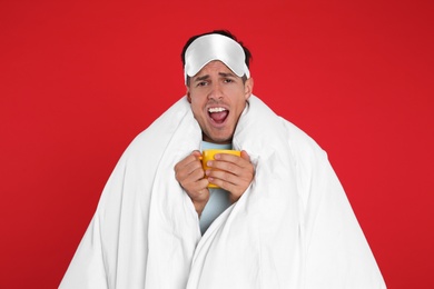 Photo of Man in sleeping mask wrapped with blanket holding cup on red background