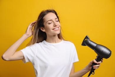 Beautiful young woman using hair dryer on yellow background