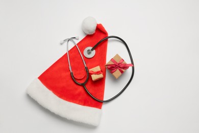 Photo of Greeting card for doctor with stethoscope, gift boxes and Santa hat on white background, top view. Space for text