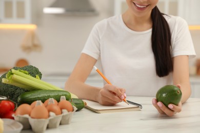 Photo of Happy woman writing in notebook near products at table, closeup. Keto diet