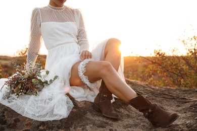 Photo of Beautiful bride with field bouquet sitting on rock at sunset, closeup