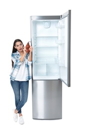 Young woman with no money in wallet near empty refrigerator on white background