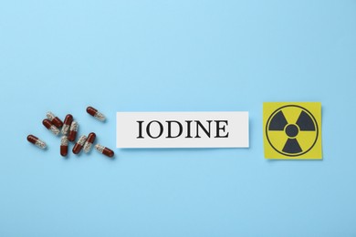 Paper note with word Iodine, radiation sign and pills on light blue background, flat lay