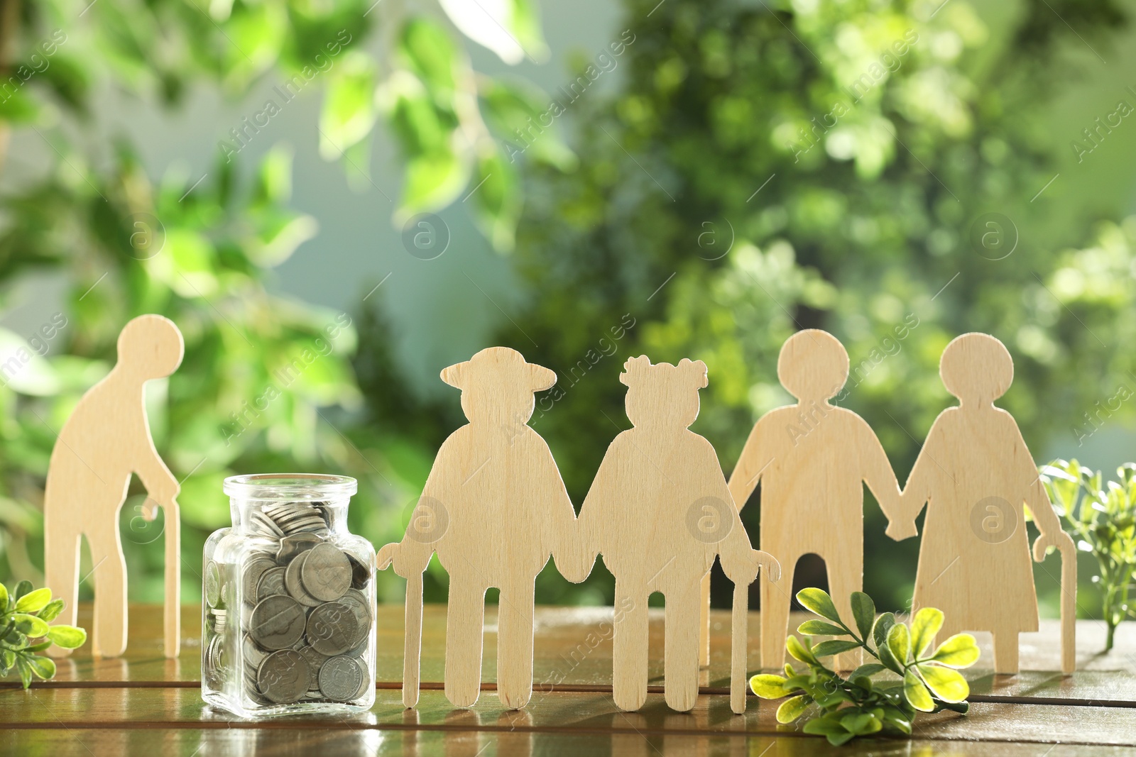 Photo of Pension savings. Figures of elderly people, coins in jar and green twigs on wooden table outdoors