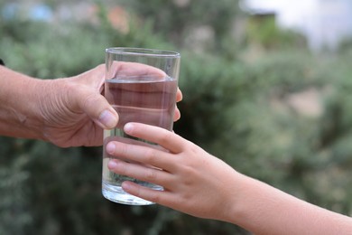 Photo of Child giving glass of water to elderly woman outdoors, closeup