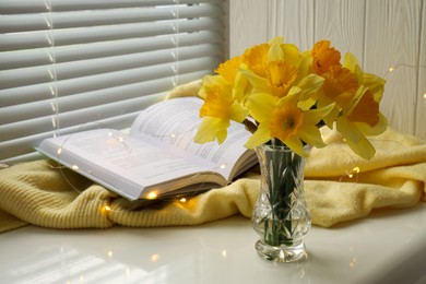 Photo of Beautiful yellow daffodils in vase, book and festive lights on windowsill. Space for text