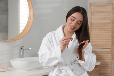 Photo of Happy young woman applying essential oil onto hair in bathroom