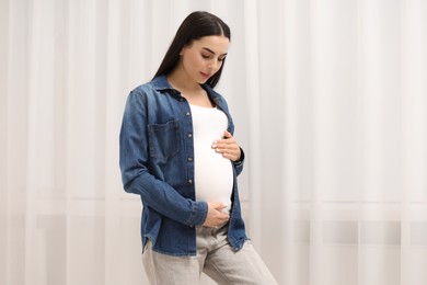 Photo of Pregnant young woman touching her belly near window indoors