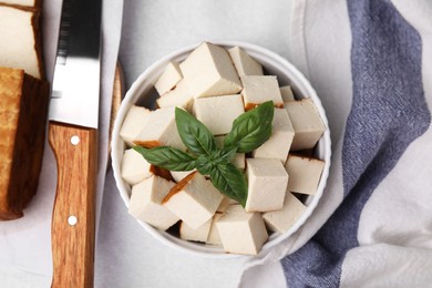 Photo of Bowl of smoked tofu cubes with basil and knife on white table, flat lay