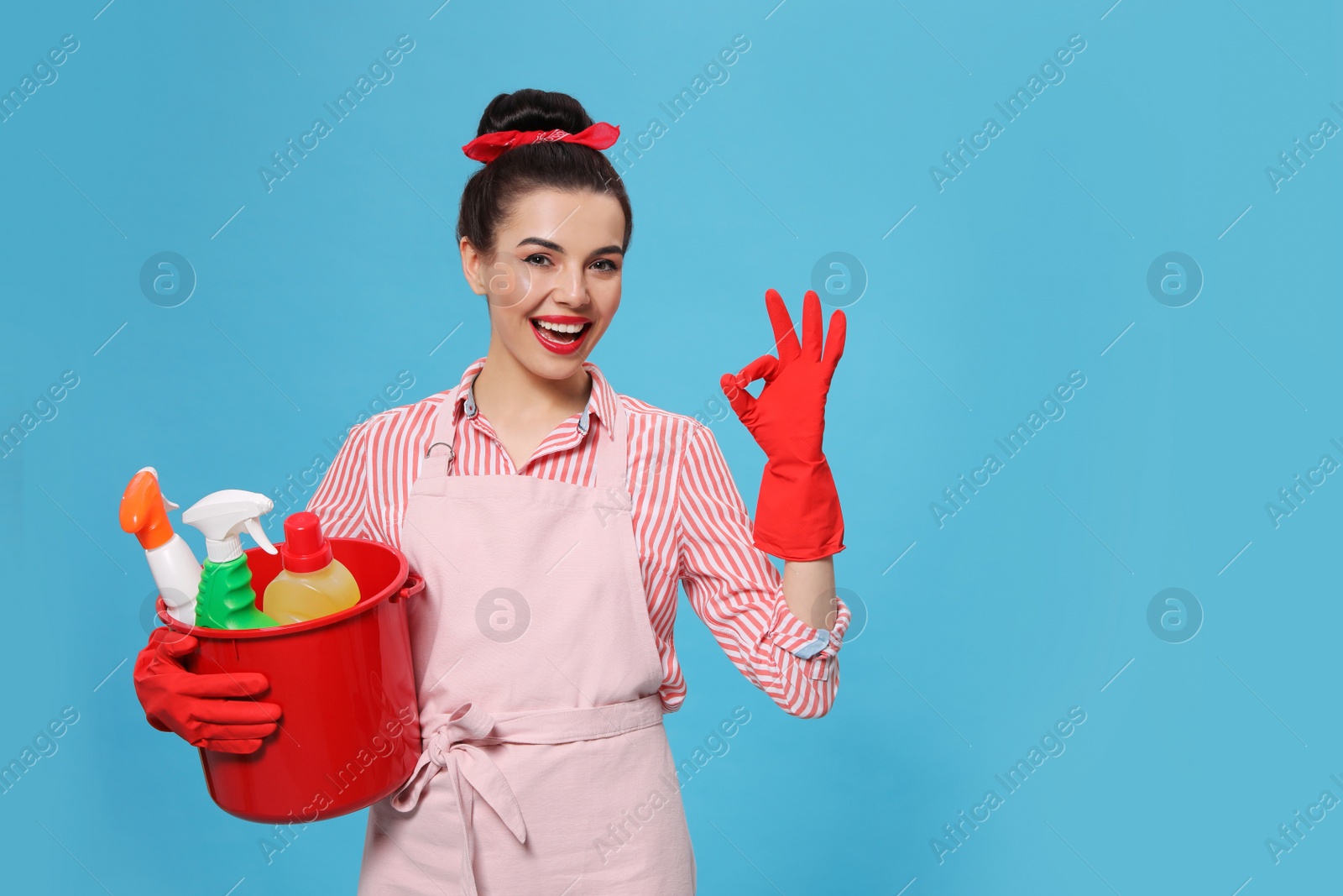 Photo of Young housewife holding bucket with cleaning supplies on light blue background, space for text