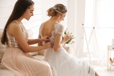 Photo of Young woman helping bride to put on wedding dress in room