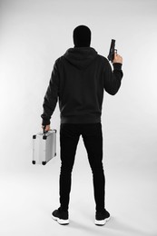 Photo of Man wearing black balaclava with metal briefcase and gun on light grey background, back view