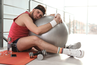 Photo of Lazy young man with sport equipment on yoga mat indoors