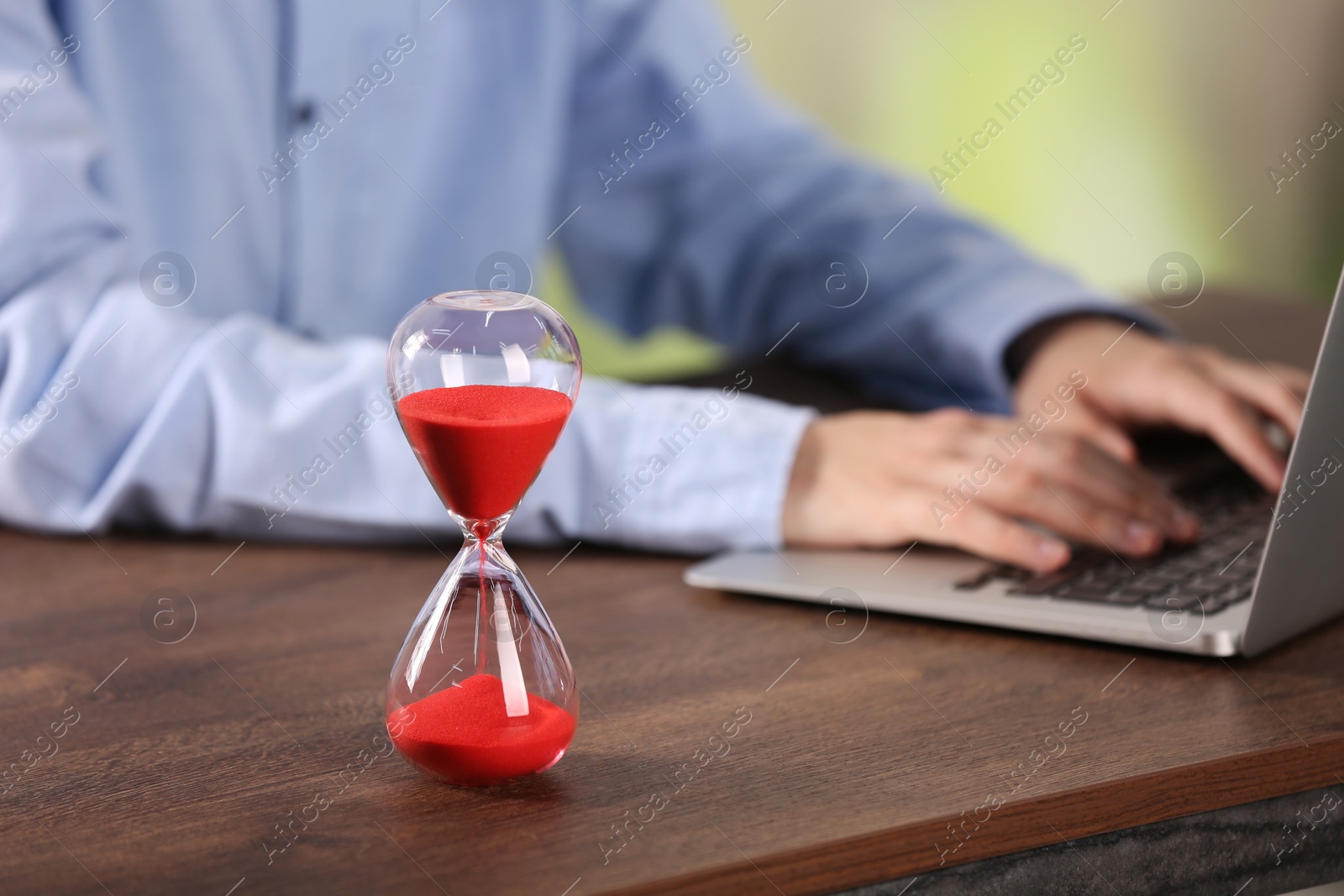 Photo of Hourglass with flowing sand on wooden table, selective focus. Man using laptop indoors