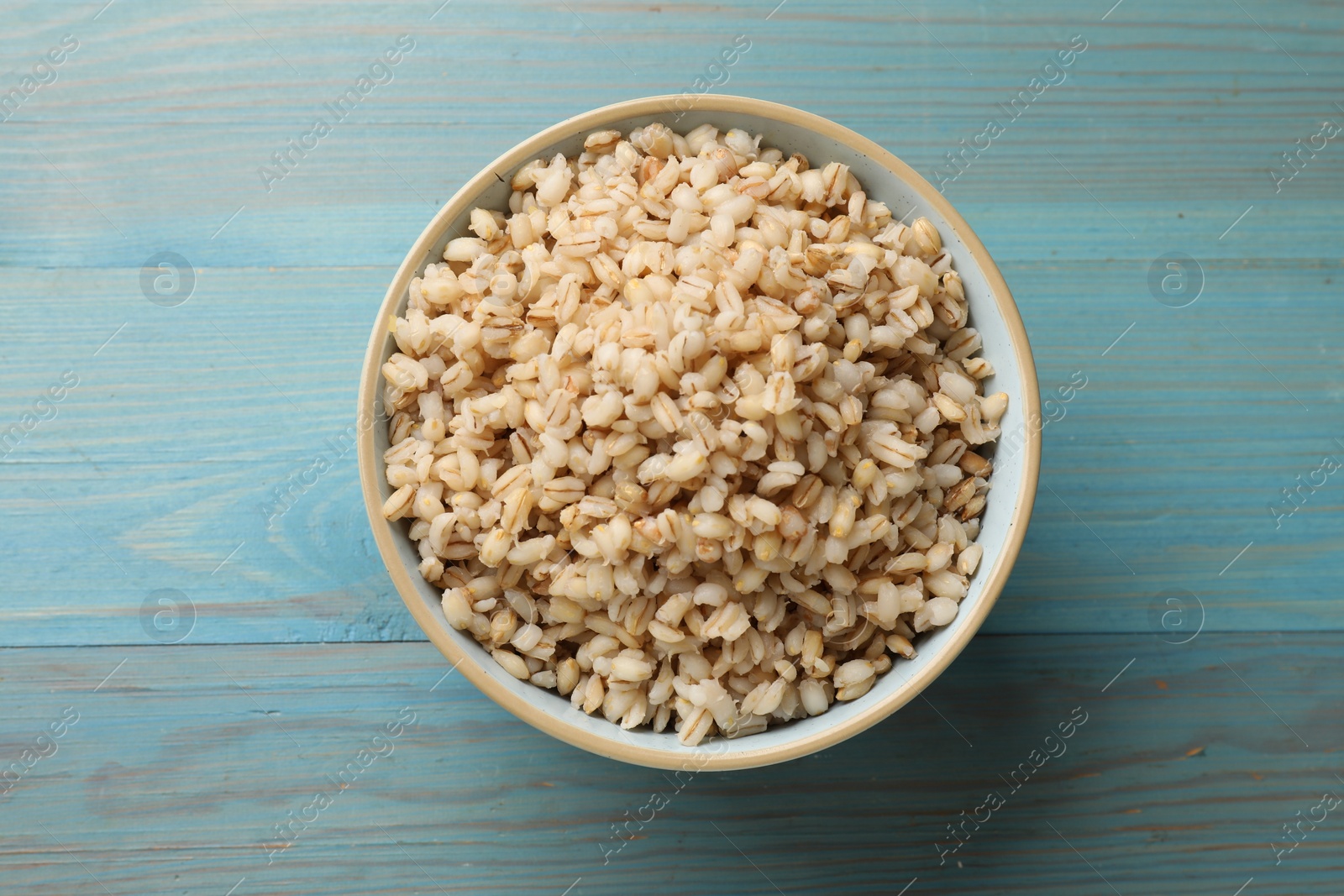 Photo of Delicious pearl barley in bowl on light blue wooden table, top view