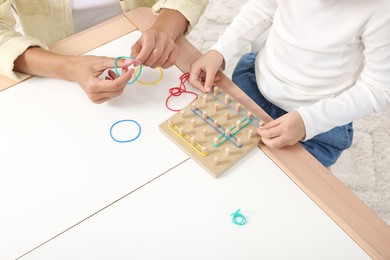 Photo of Motor skills development. Mother helping her daughter to play with geoboard and rubber bands at white table, closeup
