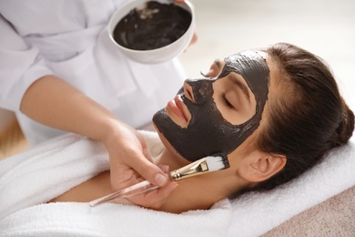 Cosmetologist applying black mask onto woman's face in spa salon