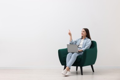 Beautiful woman with laptop pointing at something while sitting in armchair near white wall indoors, space for text