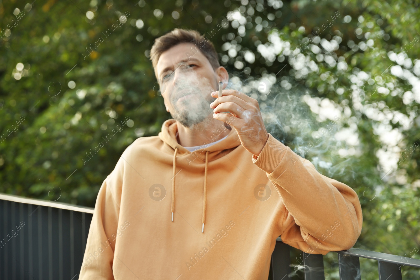 Photo of Man smoking cigarette near railing outdoors. Bad habit