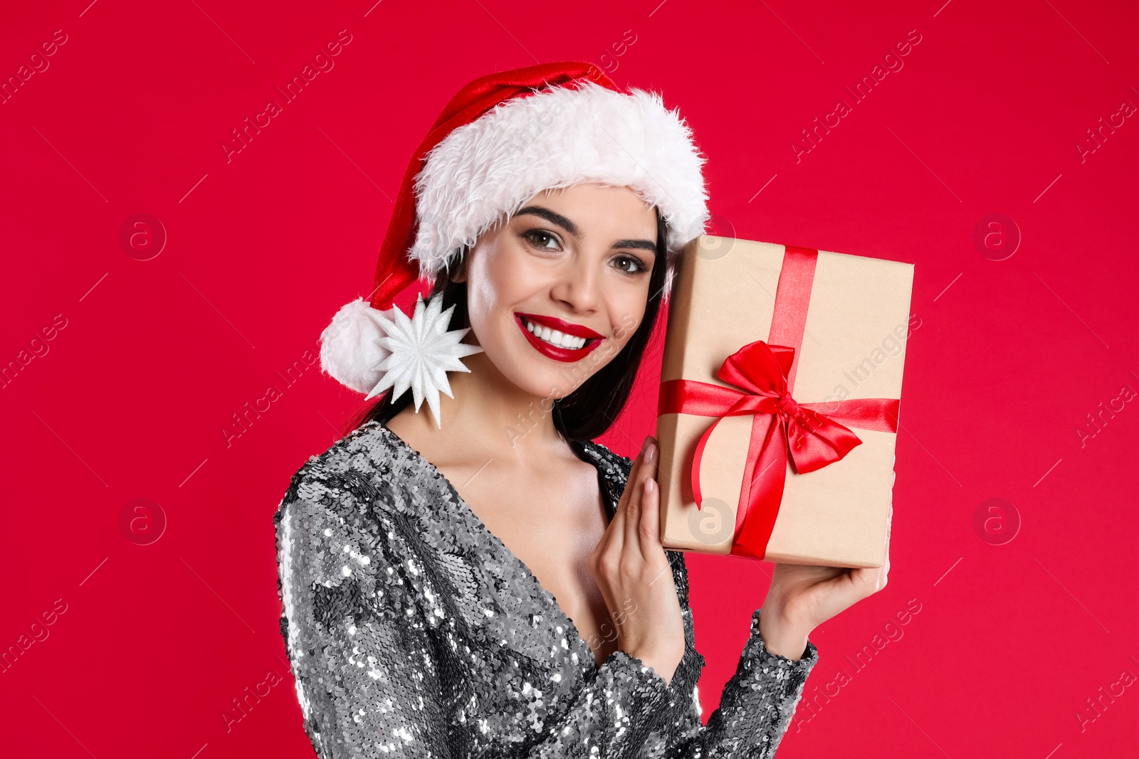 Photo of Woman in silver dress and Santa hat holding Christmas gift on red background