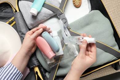 Woman putting small bottle with personal care product into plastic bag near suitcase, closeup. Cosmetic travel kit