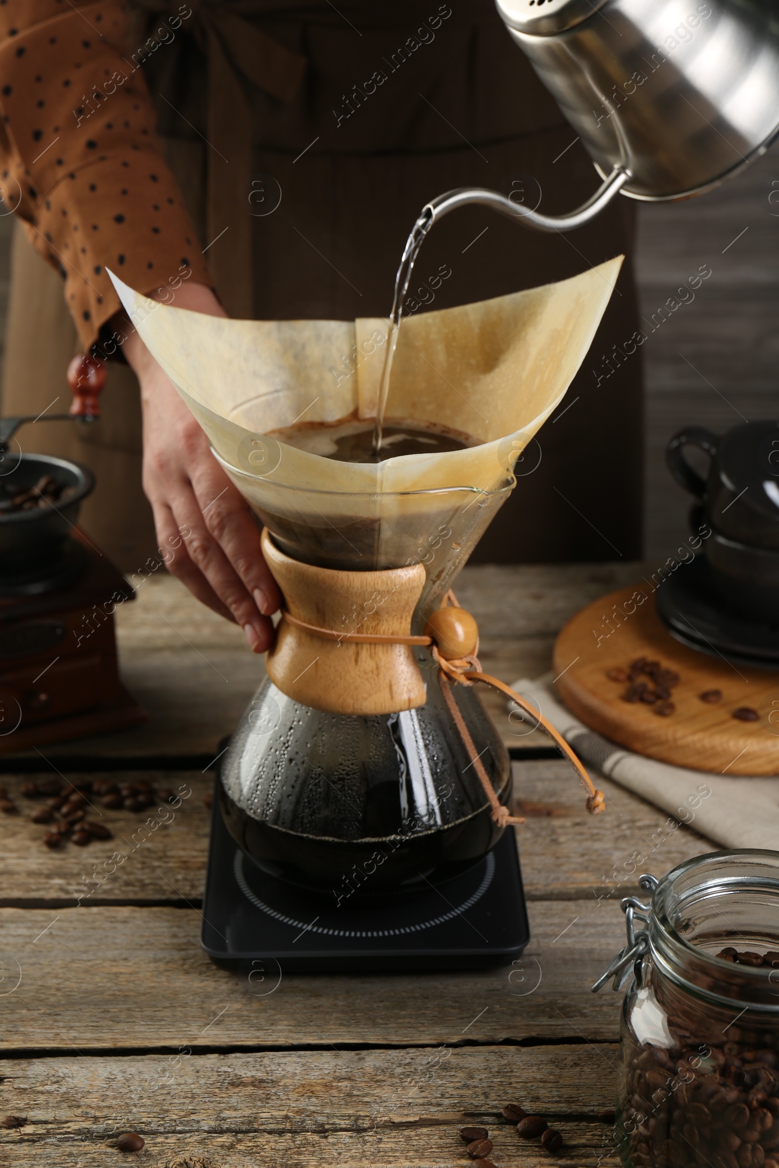 Photo of Woman pouring hot water into glass chemex coffeemaker with paper filter at wooden table, closeup