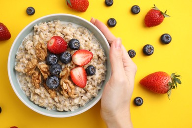 Photo of Woman holding bowl of tasty oatmeal with strawberries, blueberries and walnuts on yellow background, top view