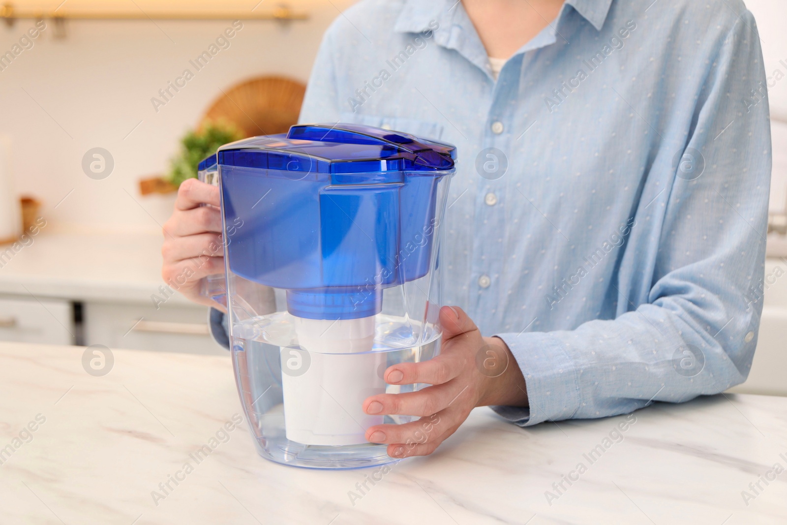 Photo of Woman with water filter jug at white marble table in kitchen, closeup