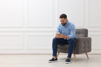 Photo of Handsome man using smartphone while sitting in armchair near white wall indoors, space for text