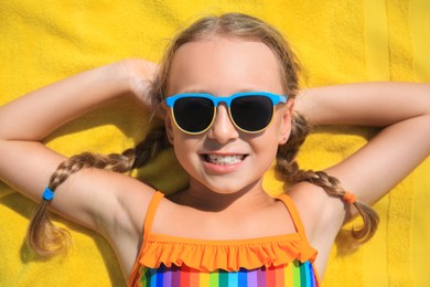 Happy little girl in sunglasses lying on beach towel outdoors, top view