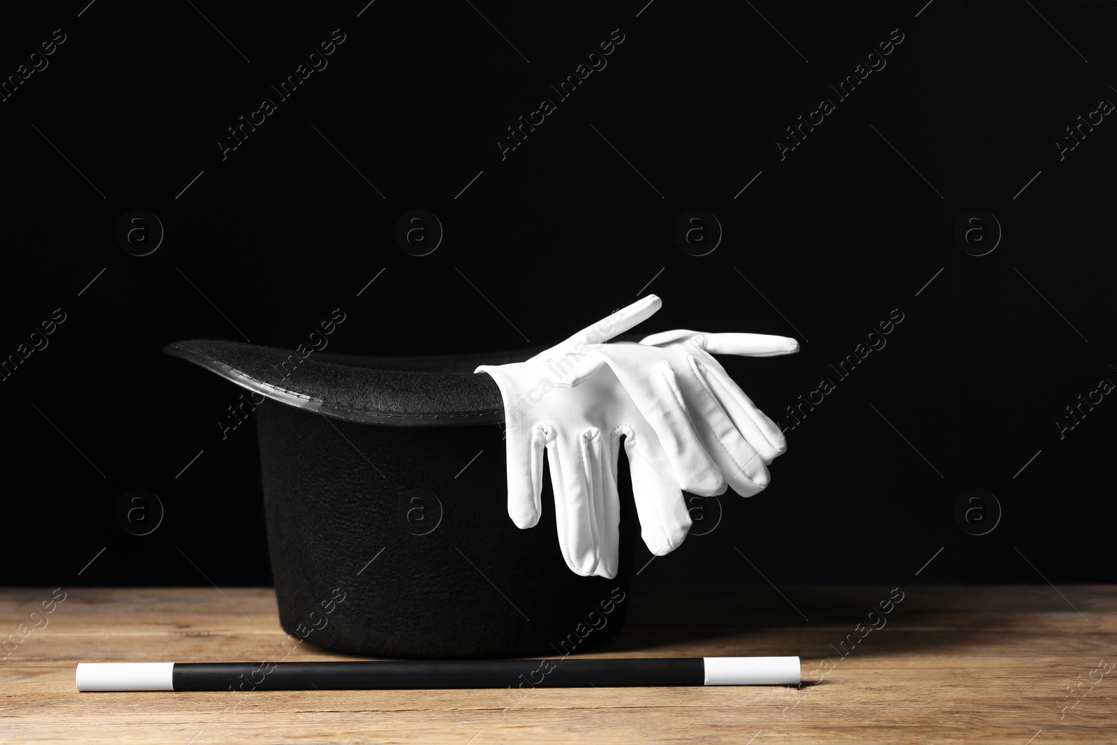 Photo of Magician's hat, gloves and wand on wooden table against black background