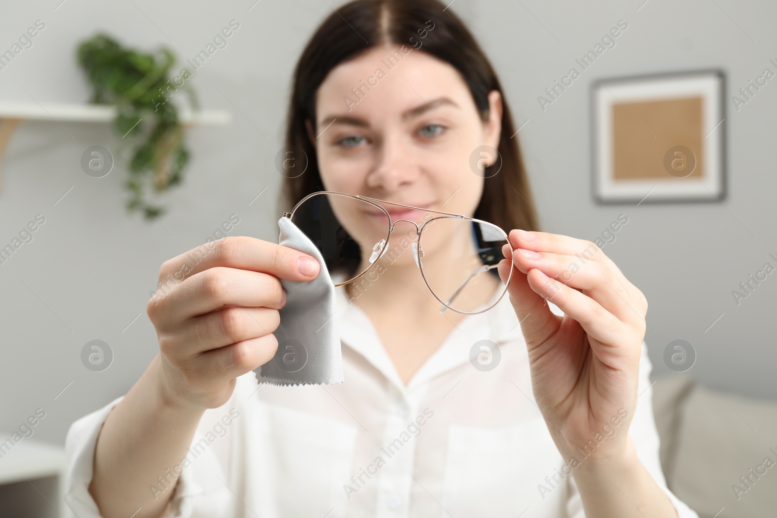 Photo of Beautiful woman cleaning glasses with microfiber cloth indoors, selective focus