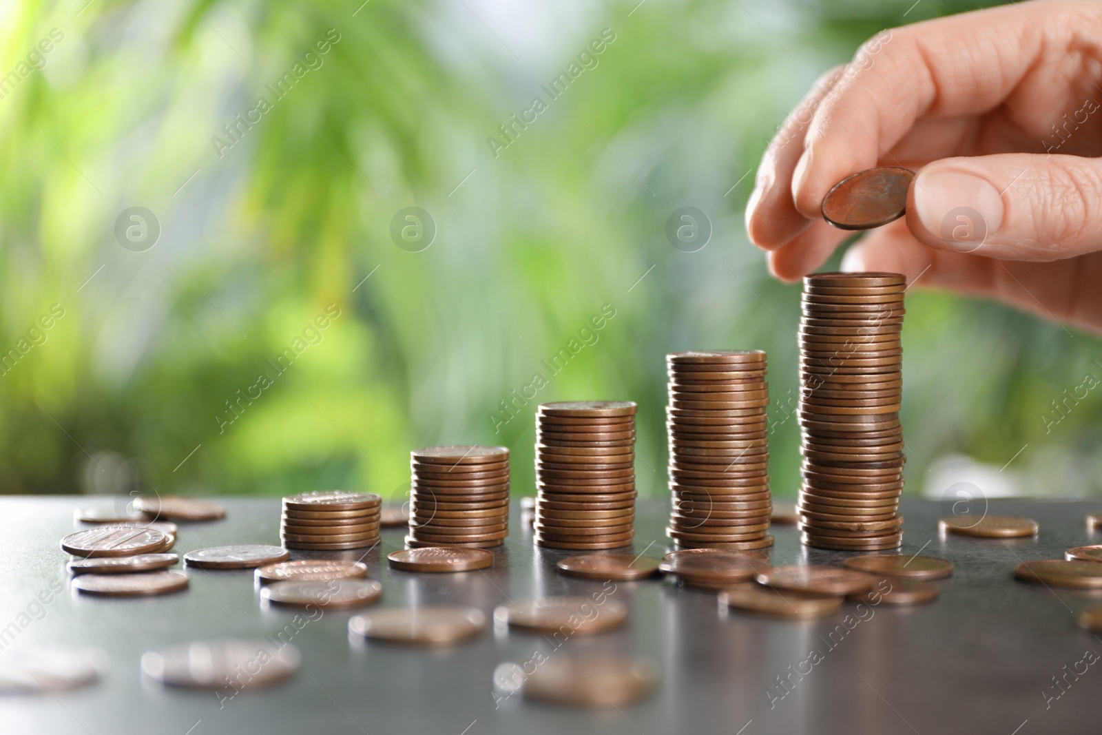 Photo of Woman stacking coins at grey table against blurred green background, closeup. Space for text