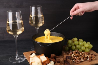 Woman dipping piece of bread into fondue pot with melted cheese at black wooden table, closeup