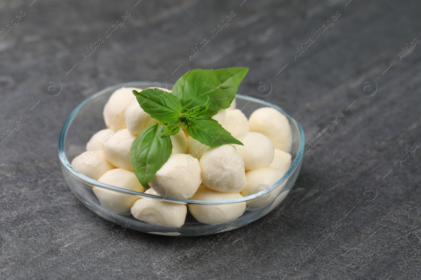Photo of Tasty mozzarella balls and basil leaves in bowl on grey table