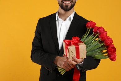 Photo of Happy man with red tulip bouquet and gift box on yellow background, closeup. 8th of March celebration