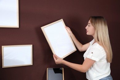 Young woman hanging empty frame on brown wall indoors. Mockup for design